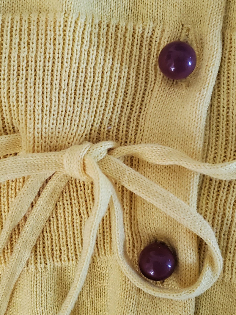 Puede incluir: Primer plano de un c&aacute;rdigan de punto amarillo p&aacute;lido. El c&aacute;rdigan tiene un cierre de lazo y dos botones de color p&uacute;rpura oscuro. El tejido de punto tiene una textura acanalada. La corbata est&aacute; hecha del mismo material que el c&aacute;rdigan.