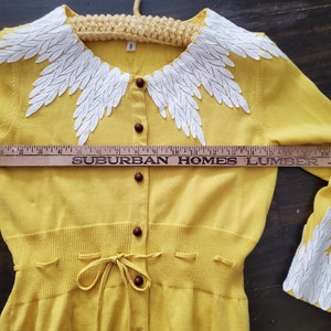 Puede incluir: C&aacute;rdigan amarillo con cuello y pu&ntilde;os bordados con motivos blancos en forma de hojas. El su&eacute;ter tiene botones marrones y un lazo en la cintura. El texto "SUBURBAN HOMES LUMBER" es visible en una regla.