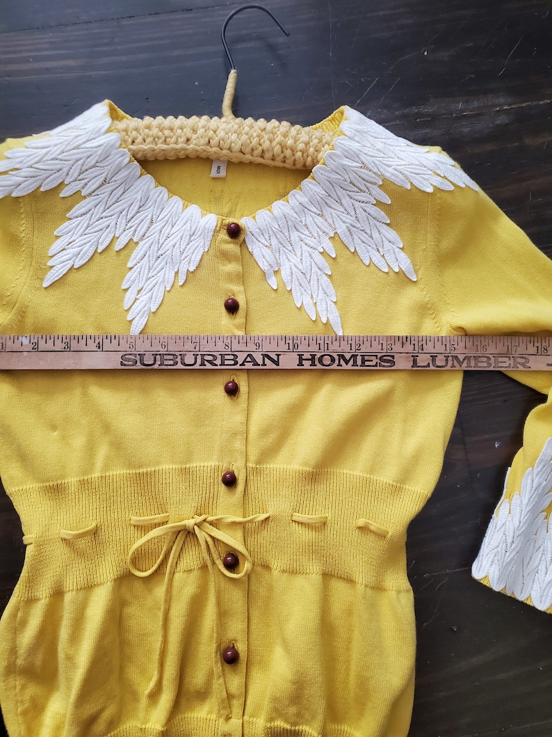 Puede incluir: C&aacute;rdigan amarillo con cuello y pu&ntilde;os bordados con motivos blancos en forma de hojas. El su&eacute;ter tiene botones marrones y un lazo en la cintura. El texto "SUBURBAN HOMES LUMBER" es visible en una regla.