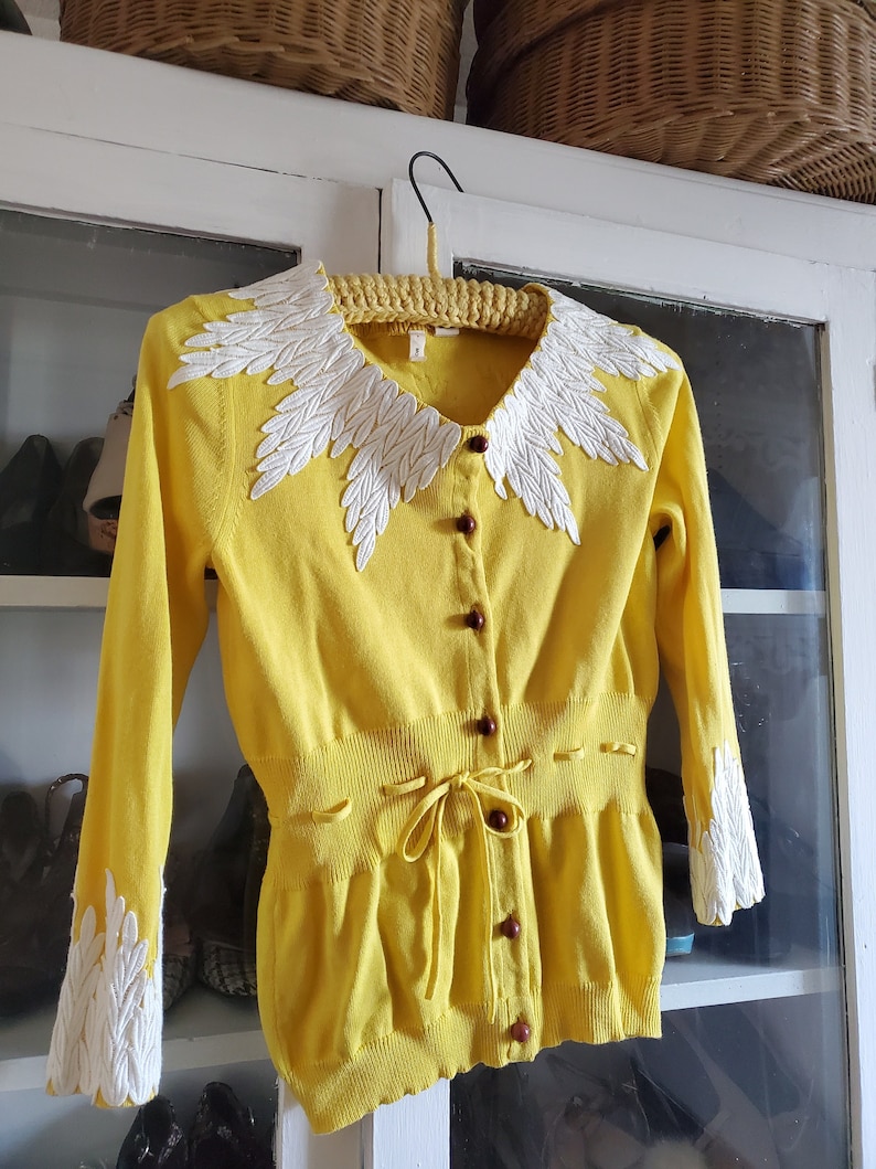 Puede incluir: C&aacute;rdigan amarillo con dise&ntilde;o de hojas blancas en el cuello y pu&ntilde;os. Cuenta con botones marrones y un lazo en la cintura. La prenda est&aacute; colgada en una percha amarilla.