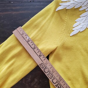 Puede incluir: Un su&eacute;ter amarillo con detalles de encaje blanco en forma de hojas en los hombros. Una regla de madera est&aacute; colocada sobre el su&eacute;ter, midiendo aproximadamente 23 cm.