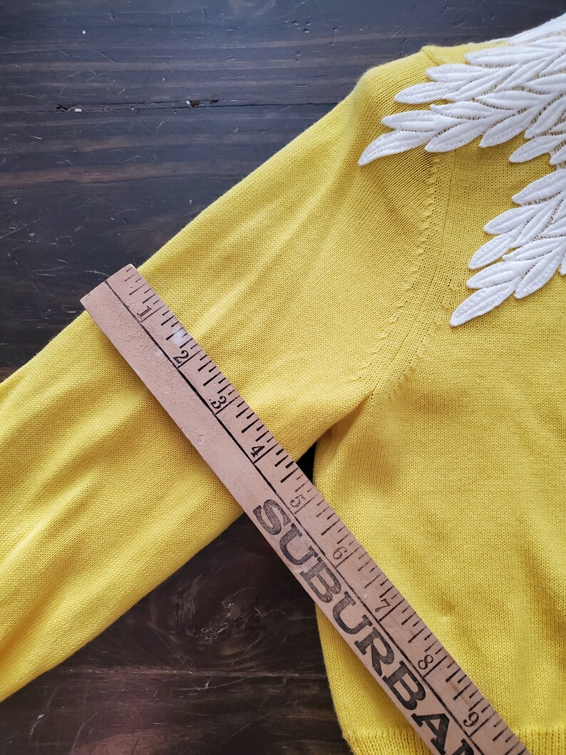 Puede incluir: Un su&eacute;ter amarillo con detalles de encaje blanco en forma de hojas en los hombros. Una regla de madera est&aacute; colocada sobre el su&eacute;ter, midiendo aproximadamente 23 cm.