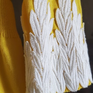 Puede incluir: Primer plano de una manga de su&eacute;ter amarilla con detalles blancos bordados en forma de hojas. El bordado blanco est&aacute; en el pu&ntilde;o de la manga. El su&eacute;ter es de color amarillo brillante.
