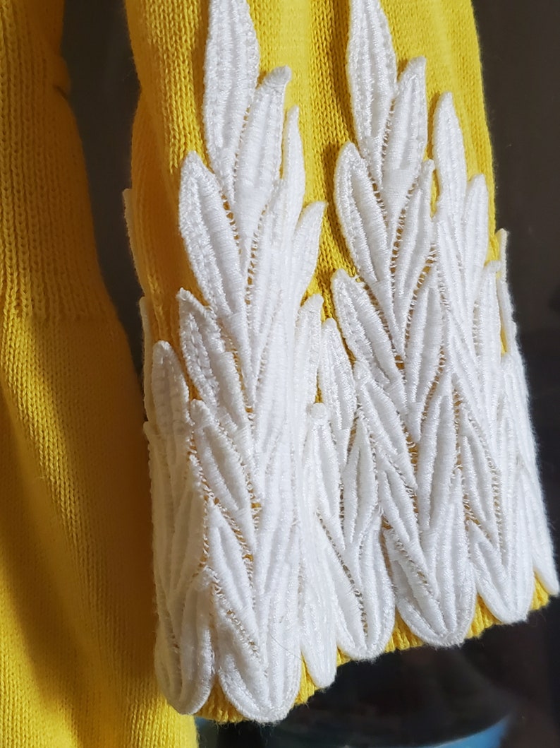 Puede incluir: Primer plano de una manga de su&eacute;ter amarilla con detalles blancos bordados en forma de hojas. El bordado blanco est&aacute; en el pu&ntilde;o de la manga. El su&eacute;ter es de color amarillo brillante.