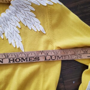 Puede incluir: Un su&eacute;ter amarillo con bordados blancos en forma de hojas en el cuello y las mangas. El su&eacute;ter se mide con una regla de madera, mostrando una longitud de aproximadamente 38 cm.