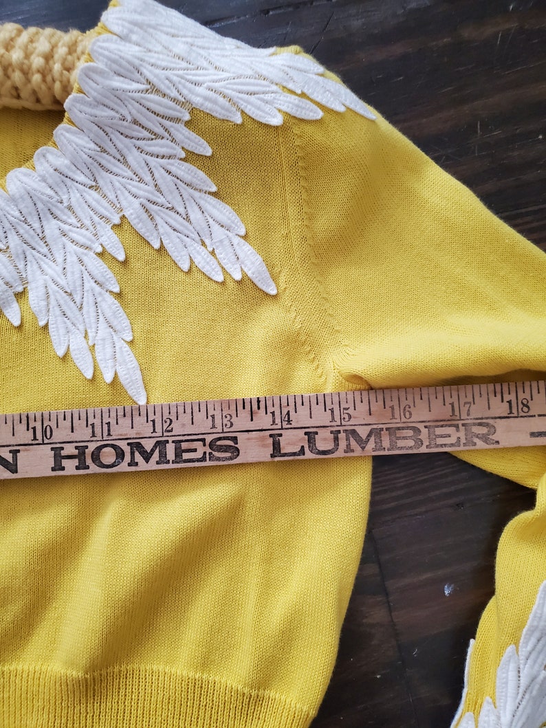Puede incluir: Un su&eacute;ter amarillo con bordados blancos en forma de hojas en el cuello y las mangas. El su&eacute;ter se mide con una regla de madera, mostrando una longitud de aproximadamente 38 cm.