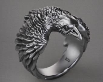 May include: A silver ring featuring a detailed design of two eagle heads facing each other. The ring is crafted from metal and has a smooth, polished finish.