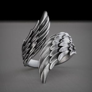 Pu&ograve; includere: Un anello color argento con un design ad ali dettagliato. L'anello presenta due ali che si avvolgono attorno al dito, con ogni ala che mostra intricati dettagli di piume. L'anello &egrave; posto su uno sfondo grigio.