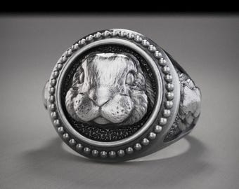 May include: A silver-toned ring featuring a detailed rabbit face within a circular frame. The ring has a textured band and a beaded border around the rabbit's head. The rabbit's face is intricately carved, showcasing its features.