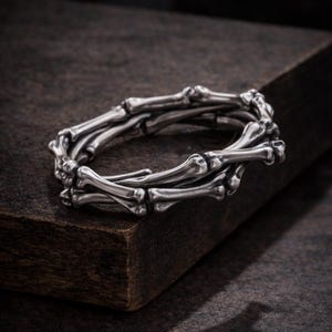 Puede incluir: Un brazalete plateado con diseño de huesos. El brazalete está hecho de múltiples eslabones con forma de hueso, creando un accesorio único y atrevido. Se exhibe sobre una superficie de madera oscura, resaltando su brillo metálico.