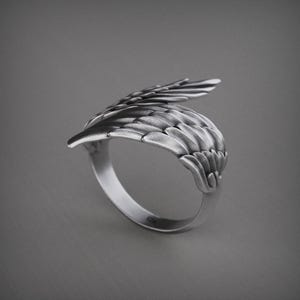 Pu&ograve; includere: Un anello color argento con un design ad ala dettagliato. L'anello presenta una forma ad ala curva con dettagli di piume individuali, creando un aspetto strutturato. L'anello &egrave; posto su uno sfondo grigio.