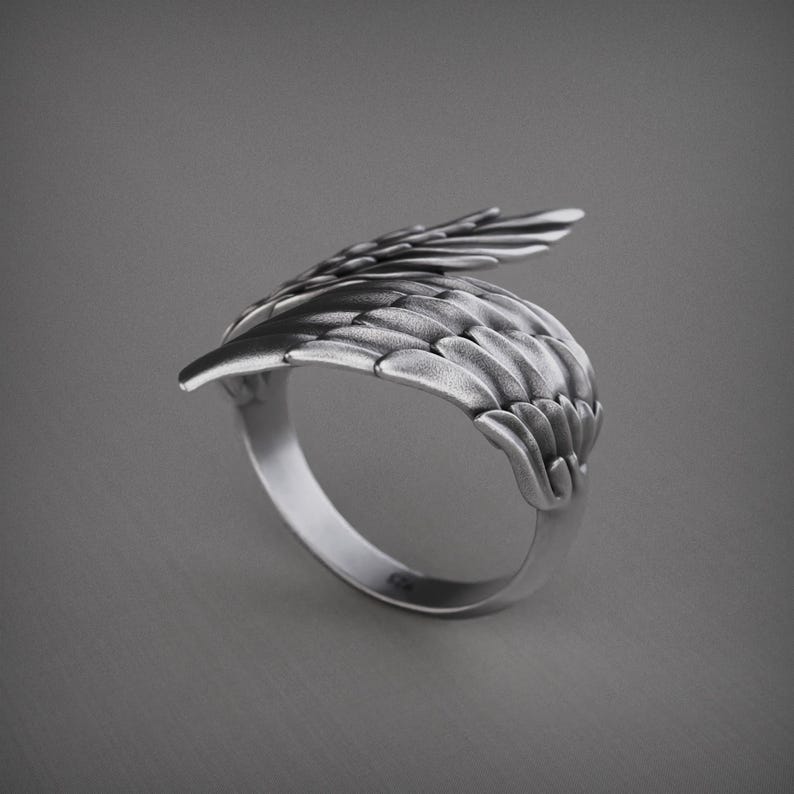 Pu&ograve; includere: Un anello color argento con un design ad ala dettagliato. L'anello presenta una forma ad ala curva con dettagli di piume individuali, creando un aspetto strutturato. L'anello &egrave; posto su uno sfondo grigio.