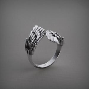 Pu&ograve; includere: Un anello color argento con un design aperto. L'anello presenta due sezioni ornate e strutturate che sembrano piume o foglie stilizzate. L'anello &egrave; posto su uno sfondo grigio.