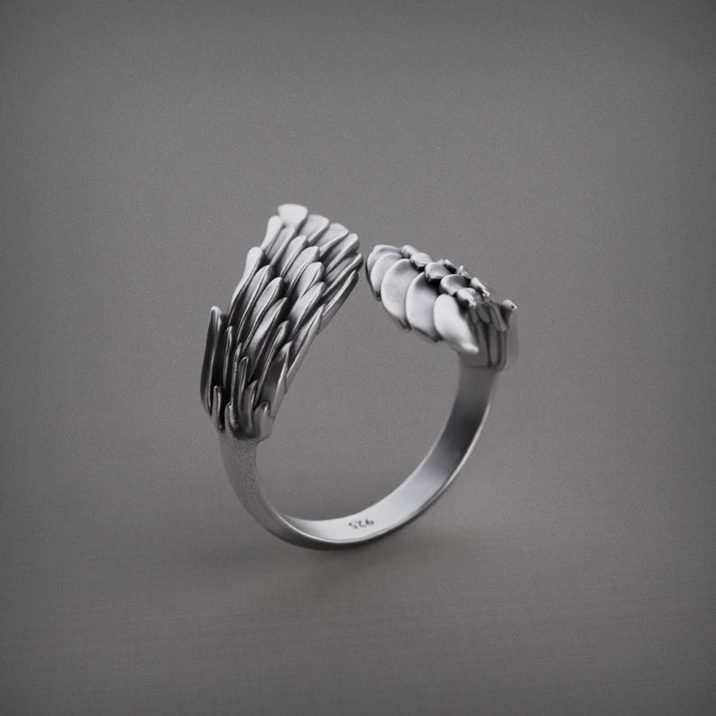 Pu&ograve; includere: Un anello color argento con un design aperto. L'anello presenta due sezioni ornate e strutturate che sembrano piume o foglie stilizzate. L'anello &egrave; posto su uno sfondo grigio.