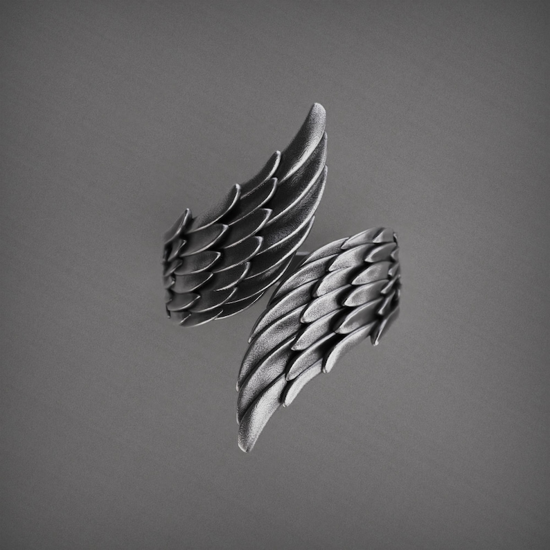 Pu&ograve; includere: Un anello argentato con un design ad ali. L'anello presenta due ali dettagliate, ciascuna con dettagli di piume a strati. Le ali sono posizionate per avvolgere un dito. L'anello &egrave; posto su uno sfondo grigio.