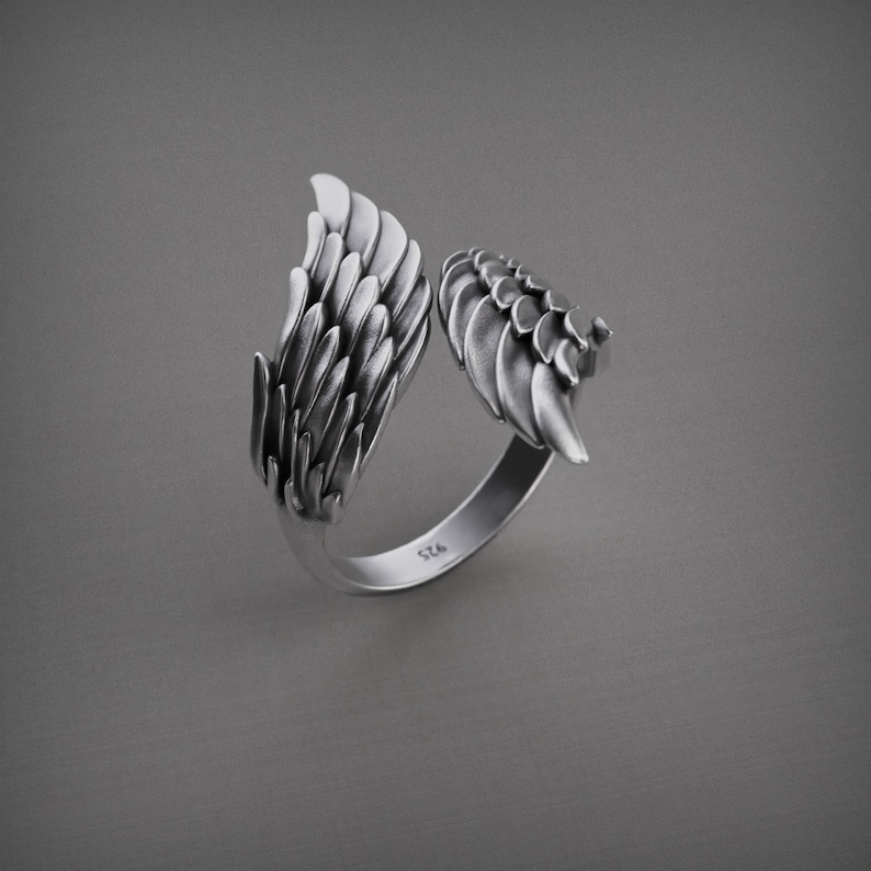 May include: A silver ring with a wing design. The ring is made of two wings that are joined together at the base. The wings are detailed with individual feathers. The ring is shown on a gray background.