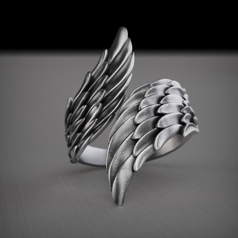 Pu&ograve; includere: Un anello color argento con un design ad ali dettagliato. L'anello presenta due ali che si avvolgono attorno al dito, con ogni ala che mostra intricati dettagli di piume. L'anello &egrave; posto su uno sfondo grigio.
