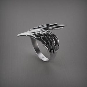 Pu&ograve; includere: Un anello d'argento con un design ad ala dettagliato. L'anello presenta una fascia curva e un'ala che avvolge il dito, con dettagli di piume individuali. L'anello &egrave; posto su uno sfondo grigio.