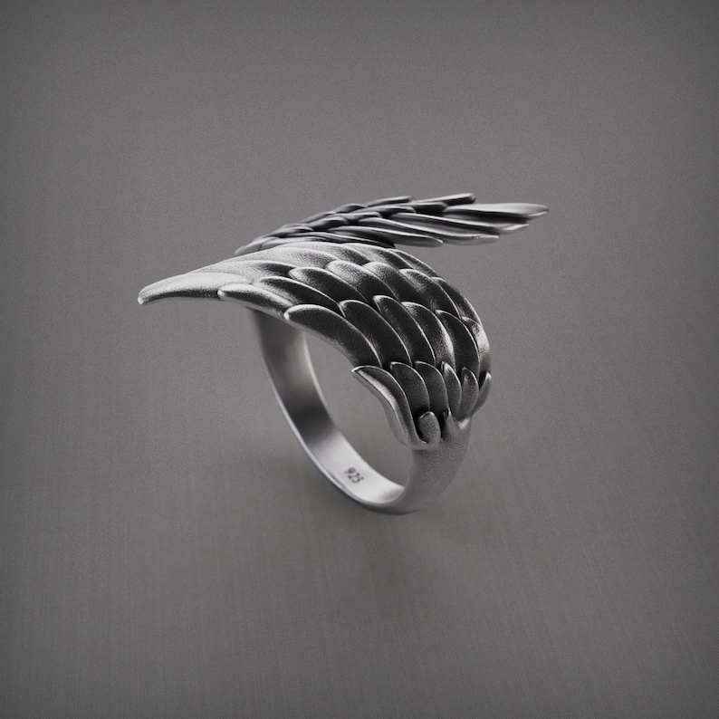 Pu&ograve; includere: Un anello d'argento con un design ad ala dettagliato. L'anello presenta una fascia curva e un'ala che avvolge il dito, con dettagli di piume individuali. L'anello &egrave; posto su uno sfondo grigio.
