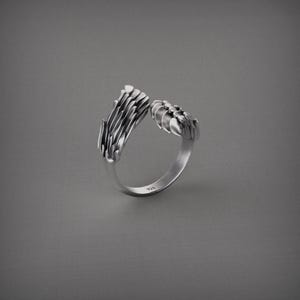 Pu&ograve; includere: Un anello color argento con un design aperto. L'anello presenta due elementi strutturati dall'aspetto organico che sembrano rami o artigli stilizzati. L'anello &egrave; posto su uno sfondo grigio.