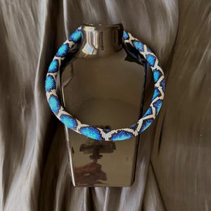 Könnte beinhalten: Eine Halskette aus Perlen mit einem geometrischen Muster in Blau, Weiß und Schwarz. Die Halskette wird auf einer schwarzen Schaufensterpuppe vor grauem Hintergrund präsentiert. Die Halskette ist im Choker-Stil.