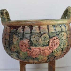 May include: A decorative ceramic basket planter with two handles. The planter features a brick-like pattern with embossed roses and leaves in shades of pink, green, and brown. The planter is supported by a wooden stand.
