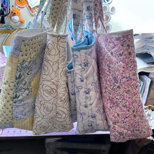 May include: A collection of quilted tote bags with floral and patterned designs. The bags feature various colors, including yellow, white, blue, and pink. Some bags have rose patterns, while others have bow and floral prints. The bags are hanging from their straps.