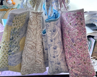 May include: A collection of quilted tote bags with floral and patterned designs. The bags feature various colors, including yellow, white, blue, and pink. Some bags have rose patterns, while others have bow and floral prints. The bags are hanging from their straps.