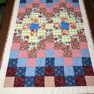 May include: A handmade patchwork quilt featuring a checkerboard pattern of various floral and solid fabrics in shades of pink, blue, red, and green. The quilt is bordered with a white fabric and a floral patterned trim.
