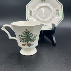 May include: A white teacup and saucer set with a Christmas tree design. The teacup has a handle and a hexagonal base. The saucer is octagonal with a green border and holly accents. The teacup and saucer are on a black surface.