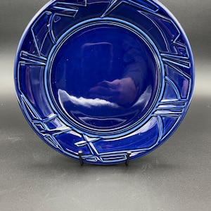 May include: A cobalt blue ceramic plate with a geometric design around the rim. The plate has a glossy finish and a slightly raised edge. The design consists of angular lines and shapes, creating a modern aesthetic. This decorative plate is suitable for serving food.