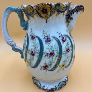 May include: A white porcelain pitcher with a floral design. The pitcher features a curved handle and spout, with teal and gold accents. The body is decorated with colorful flowers and teal accents.