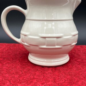 May include: A cream-colored ceramic pitcher with a handle and spout. The pitcher features a textured, basket-weave design around the body and a rounded base. The pitcher is set against a red patterned background.