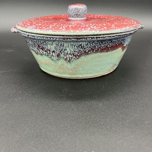May include: A ceramic bowl with a lid. The bowl is a mix of colors, including red, green, and blue. The lid is red with white speckles and a small knob. The bowl has a unique, handcrafted appearance.
