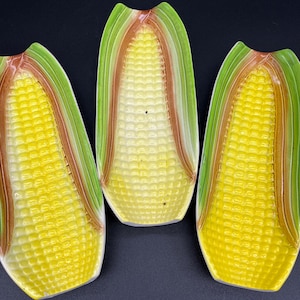 May include: Three ceramic corn-shaped serving dishes. Each dish features a yellow corn cob design with green and brown accents, mimicking the appearance of real corn. The dishes are arranged against a dark background.