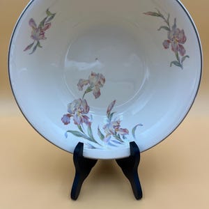 May include: A white ceramic serving bowl with a silver rim and a floral iris design in shades of purple, pink, and green. The bowl is displayed on a black stand.