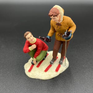 May include: A figurine set of two people in winter clothing. The person in the brown jacket is holding a bottle of Coca-Cola and a ski pole. The person in the red sweater is crouching down. The figurine set is on a white base with red stripes.
