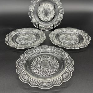 May include: A set of four clear glass plates with a decorative, scalloped edge and a central, textured design. One plate is displayed on a black stand. The plates are arranged on a black surface.