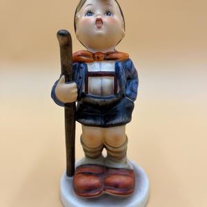 May include: A vintage porcelain figurine of a young boy in traditional attire, holding a wooden staff. The boy wears a blue jacket, dark shorts, and brown boots. The figurine has a white base and is painted with detailed features.