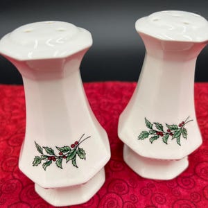 May include: A pair of white ceramic salt and pepper shakers. Each shaker has a hexagonal base and a flared top with several holes. A sprig of green leaves and red berries decorates the lower portion of each shaker. The background is a red patterned fabric.