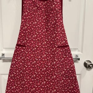 May include: A red floral apron with a dark red trim and two pockets. The apron is hanging on a black hanger against a white door. The apron is covered in small white and pink flowers.