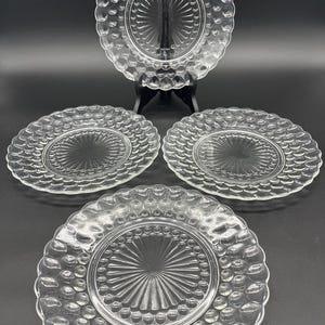 May include: Four clear glass plates with a textured, bubble-like rim and a sunburst design in the center. One plate is displayed on a black stand, while the other three are arranged below. The plates are round and appear to be vintage.