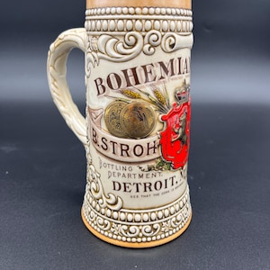 May include: Cream-colored ceramic beer stein with a handle, featuring embossed floral and scroll designs. The stein has the words "BOHEMIA" and "B. STROH BOTTLING DEPARTMENT DETROIT" printed on it, along with a red crest and gold coins.
