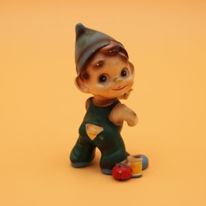 May include: A vintage ceramic figurine of a smiling gnome wearing a blue hat and green overalls. The gnome is holding a paintbrush and is next to a red tomato and two paint cans. The figurine has a whimsical, antique aesthetic.