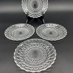 May include: A set of four clear glass plates. Each plate has a scalloped edge and a central design of radiating lines. One plate is displayed on a black stand, with the other three plates arranged in front.
