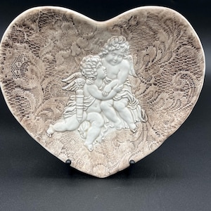May include: Heart-shaped decorative dish with a lace-like pattern in shades of brown and white. Two cherubic figures are in the center, embracing. The dish is made of ceramic or porcelain and is displayed on a black stand.