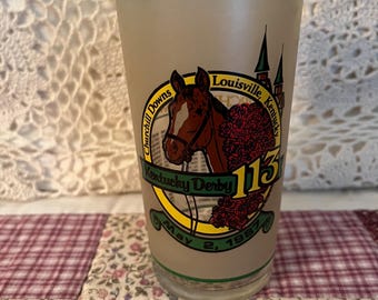 Puede incluir: Un vaso vintage con el logotipo de Kentucky Derby 113, una ilustración de un caballo, las palabras "Churchill Downs" y "Louisville, Kentucky". El vaso tiene un fondo beige con detalles verdes y dorados, y la fecha "May 2, 1987".