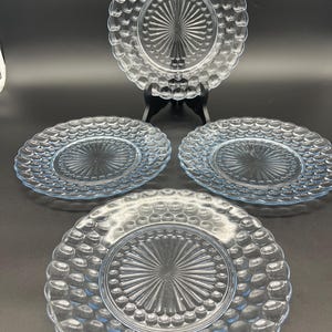 May include: Four clear glass plates with a textured, bubble-like rim and a starburst design in the center. One plate is displayed on a black stand, while the other three are arranged in front. The plates are a light blue color.