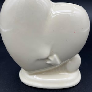 May include: Cream-colored ceramic heart-shaped vase with a rounded base. The vase has a smooth, glossy finish and a slot at the top. The heart shape is slightly asymmetrical, with a small, rounded detail on the side.