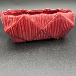 May include: A rectangular, red ceramic planter with a textured, geometric design. The planter has a wavy top edge and a series of diamond-shaped patterns. The planter is suitable for small plants or succulents.